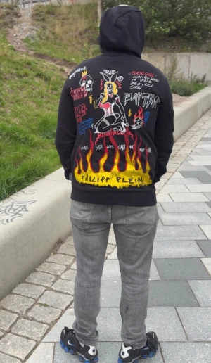 Svart Philipp Plein hoodie grim reapers med tryck - Säljer en svart hoodie från Philipp Plein med modell grim reapers limiterad med färgstarka grafiska tryck både fram och bak. Motiven inkluderar eldflammor, tecknade figurer och text i olika färger. Hoodien har huva och ribbade muddar. Perfekt för dig som gillar streetwear och unika plagg. Nypris 16316kr.limiterad hoodie säljer för 3500kr