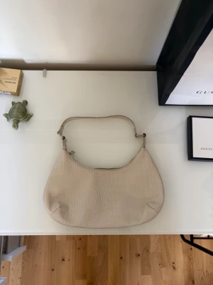Beige axelremsväska i croc-mönster - Stilren beige handväska med axelrem och croc-mönstrad yta. Väskan har en mjukt rundad form och dragkedja upptill. 
