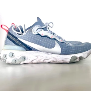 Nike React Vision grå/vit sneakers - Nike React Vision sneakers i grå och vitt med genomskinligt mesh-tyg, vita detaljer och reflexer. Sulan är vit med grå och mintgröna inslag och har en unik, vågig design. Röda dragflikar bak och stora vita swoosh-loggor på sidorna. Perfekta för dig som gillar streetwear.