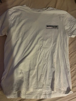 Vit Quiksilver t-shirt med tryck - Säljer en vit t-shirt från Quiksilver med tryck på bröstet och en stor färgglad print på ryggen med palmer och solnedgång. Klassisk passform och rund hals. Perfekt för dig som gillar surfvibbar och avslappnad stil.