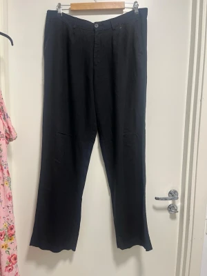 Svarta vida linnebyxor - Snygga svarta linnebyxor med raka, vida ben och klassisk midja. Byxorna har knapp och dragkedja framtill samt bälteshällor. Perfekta för en avslappnad och stilren look.