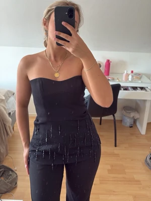 Svart axelbandslös topp med pärldetaljer - Snygg svart axelbandslös topp med figurnära passform och dekorativa pärldetaljer framtill. Toppen har en stilren och elegant look, perfekt att styla med smycken. Perfekt till något festligt eller någon utekväll . Aldrig använd. h&M edition. Nypris 500kr.