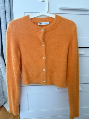 Fluffig orange kofta från Zara - Superfin fluffig orange kofta från Zara med rund halsringning och glittriga blomformade knappar. Koftan har lång ärm och är gjord i ett mjukt, lurvigt material som känns riktigt cozy. Perfekt för dig som vill sticka ut med färg och detaljer!