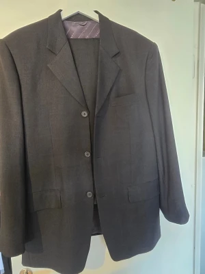 Hugo Boss, Mörkgrå klassisk kostym herr. Storlek 52 - Mörkgrå kostym med kavaj och byxor. Kavajen har två knappar framtill och fickor med lock. Insidan av kavajen har ett mönstrat foder. Byxorna matchar kavajen i färg och stil. I nyskick endast använd 1 gång.