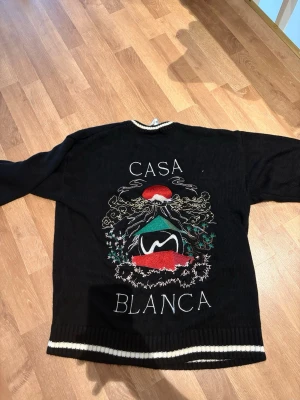 Svart stickad tröja med brodyr - Svart stickad tröja med vit kant vid hals och nederkant. Stor broderad design på ryggen med texten 'CASA BLANCA', färgglada detaljer i rött, grönt och vitt samt ett konstnärligt motiv. Perfekt för dig som vill sticka ut med en unik och trendig look.