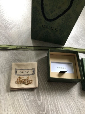 Guld GG örhängen från Gucci - Säljer ett par ikoniska örhängen från Gucci i guldfärgad metall med det klassiska dubbla G-logotypen. Kommer med originalpåse, ask och presentpåse. Perfekta för dig som vill ha en lyxig och trendig accessoar.