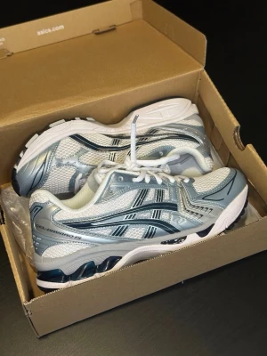 Asics gel kayano 14 - Snygga Asics sneakers i vitt, ljusblått och silver med mesh- och syntetmaterial. Skorna har klassisk låg profil, vita snören och detaljer i grått och blått. Perfekta för dig som gillar sportig och trendig stil. Sulan är rejäl och ger bra stöd.