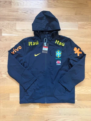 Svart Brasil vindjacka med huva - Snygg svart vindjacka med huva och dragkedja framtill. Jackan har Brasil-logga, Nike-logga och flera färgglada tryck i gult och orange på bröst och ärmar. Perfekt för dig som gillar sportig stil och vill sticka ut.