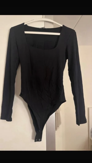 Svart långärmad body/topp med fyrkantig ringning - Snygg svart body med lång ärm och fyrkantig ringning. Tillverkad i stretchigt material som sitter tight mot kroppen. Praktisk knäppning i grenen. Perfekt att styla med jeans eller kjol för en clean look. NY UTAN PRISLAPP