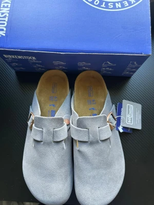 Grå mockatofflor från Birkenstock - Snygga grå tofflor från Birkenstock i mjuk mocka med klassisk slip-in design och justerbart spänne i metall. Fotbädden är ergonomisk och innersulan är i skönt material för extra komfort. Perfekta för dig som gillar stilrena och bekväma skor.