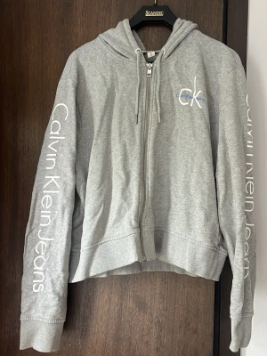 Grå hoodie från Calvin Klein Jeans - Snygg grå zip-hoodie från Calvin Klein Jeans med logga på bröstet och text längs båda ärmarna. Hoodien har dragkedja framtill, ribbade muddar och justerbar huva med snören.