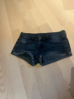 Mörkblå jeansshorts med uppvik - Snygga mörkblå jeansshorts med uppvikta benslut och klassisk femficksdesign. Shortsen har knapp och dragkedja framtill samt detaljerade sömmar. Perfekta för varma dagar och enkel att matcha med olika toppar.