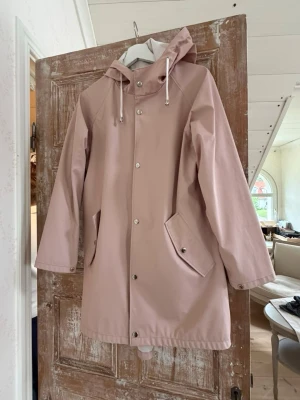 Ljusrosa regnjacka med huva - Snygg ljusrosa regnjacka med huva och dragsko, tryckknappar framtill och två snedställda fickor med lock. Jackan har raka ärmar och är tillverkad i ett vattenavvisande material som passar perfekt för regniga dagar.