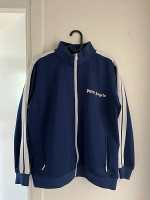 Blå zip-tröja från Palm Angels - Snygg blå zip-tröja från Palm Angels med vita ränder längs ärmarna och vit dragkedja. Tröjan har hög krage, två fickor med dragkedja och logga på bröstet. Perfekt för en sportig och trendig look.