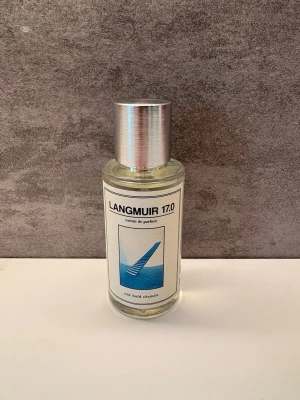 Langmuir 17.0 parfym - En stilren parfymflaska från Une Boule Chemin med namnet Langmuir 17.0. Flaskan är cylindrisk i genomskinligt glas med en silverfärgad metallkork. Etiketten har en minimalistisk design med blått motiv och svart text.