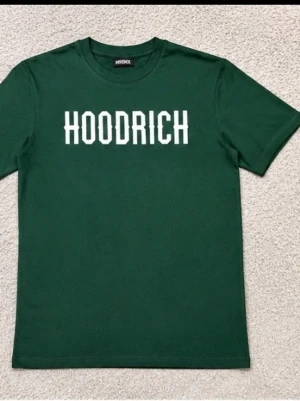 Grön HOODRICH t-shirt med tryckk - Snygg grön t-shirt från HOODRICH med vitt tryck på bröstet. Klassisk passform med rund hals och korta ärmar. Perfekt för en chill och avslappnad stil. Materialet känns mjukt och bekvämt mot huden.