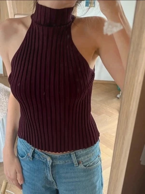 Ribbad vinröd halterneck topp - Snygg ribbad topp i vinrött med hög hals och halterneck-modell. Toppen är ärmlös och har en tight passform som framhäver figuren. Perfekt att matcha med jeans eller kjol för en trendig look. Elastisk så den passar många storlekar! Aldrig använd 
