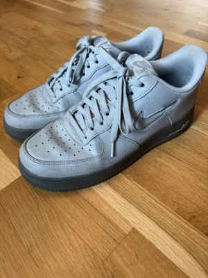 Nike Air Force 1 Low grå sneakers - Säljer ett par grå Nike Air Force 1 Low sneakers i skinn med klassisk siluett och perforerad tåbox. Skorna har matchande grå snören, Nike Swoosh på sidan och en robust sula med AIR-detalj. Perfekta för dig som gillar stilrena och tidlösa sneakers.