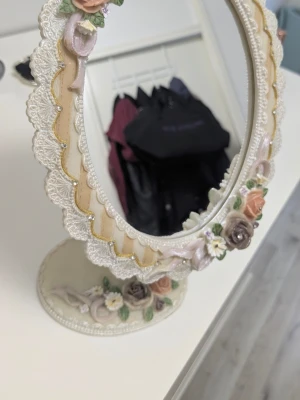 Romantisk sminkspegel med blommor - Söt oval sminkspegel på fot med dekorativ ram i krämvitt och pastellfärger. Spegeln har detaljer i spets, guld och vackra blommor i rosa, beige och lila. Perfekt för sminkbordet och ger en vintagekänsla. Materialet är tungt! troligen keramik/resin/porslin.