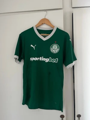 Grön Palmeiras matchtröja Puma - Snygg grön matchtröja från Palmeiras med vit V-ringning och vita detaljer. Puma-logga på bröstet och Palmeiras klubbmärke samt sponsortryck 'sportingbet' framtill. Perfekt för fotbollsfans som vill sticka ut på planen. Storlek L men mer som en M. 