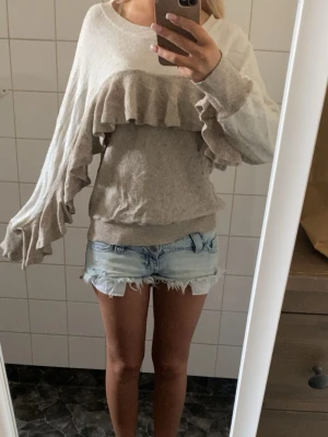 Beige stickad tröja med volanger - Supersnygg beige stickad tröja med bred volangdetalj över bröstet och längs ärmarna. Tröjan har rund halsringning, långa ärmar och ribbade muddar. Perfekt för dig som gillar en trendig och unik look.