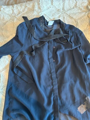 Mörkblå blus med dekorband - Snygg mörkblå blus med långa ärmar och dekorativa svarta band med glittriga detaljer vid halsen. Blusen har knäppning framtill och ett lätt, flytande tyg som ger en avslappnad men ändå chic look.