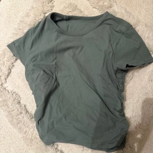 Grön ribbad t-shirt från Lululemon - Mörkgrön ribbad t-shirt från Lululemon med rund halsringning och korta ärmar. Tröjan har en tight passform och rynkade detaljer på ena sidan, vilket ger en sportig och modern look. Perfekt för träning eller en aktiv vardag.