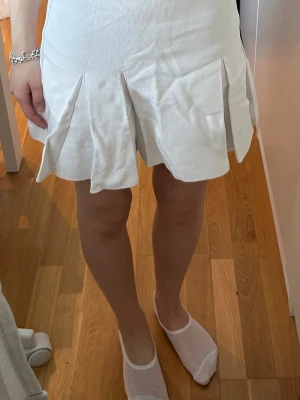 Vit kortkjol med plisserade detaljer - Snygg vit kortkjol med plisserade detaljer som ger en sportig och trendig look. Kjolen har en hög midja och är tillverkad i ett mjukt bomullsmaterial som känns bekvämt mot huden. Perfekt för dig som vill ha en fräsch och stilren kjol till sommaren.