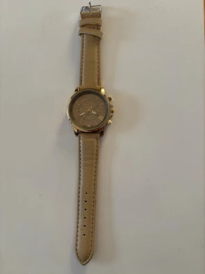 Beige klocka med gulddetaljer - Stilren klocka med beige läderarmband och guldfärgat ur. Urtavlan är också beige med flera små visare och detaljer. Perfekt accessoar för dig som gillar en klassisk och enkel look.