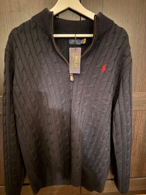 Mörkblå kabelstickad tröja Polo Ralph Lauren - Kabelstickad mörkblå tröja från Polo Ralph Lauren med halv dragkedja och röd broderad logga på bröstet. Tröjan har lång ärm och ribbade muddar. Perfekt för en stilren och klassisk look. )Pris kan diskuteras!)