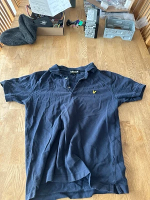 Mörkblå pikétröja från Lyle & Scott - Klassisk mörkblå pikétröja från Lyle & Scott med korta ärmar och gul broderad logga på bröstet. Tröjan har krage och knappar framtill. Perfekt för en avslappnad men ändå stilren look.