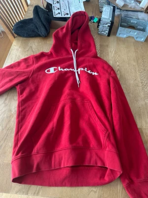 Röd Champion hoodie med logga - Snygg röd hoodie från Champion med stor vit logga över bröstet. Klassisk känguruficka framtill och justerbar huva med vita snören. Tillverkad i mjukt och bekvämt material, perfekt för en avslappnad stil.