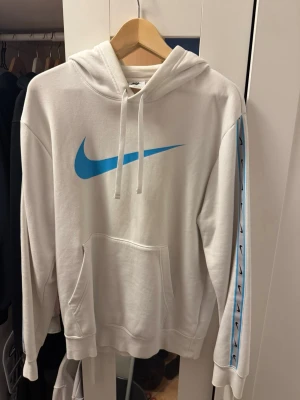 Vit Nike hoodie med blå logga - Säljer en vit hoodie från Nike med stor blå Swoosh-logga på bröstet och blå detaljer längs ärmarna. Hoodien har huva med dragsko och en stor magficka. Perfekt för dig som gillar sportig och clean stil. STORLEK S
