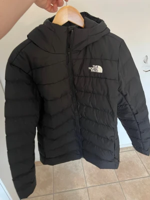 Svart dunjacka från The North Face - Säljer en svart dunjacka från The North Face med huva och dragkedja framtill. Jackan har ett diskret vitt logotryck på bröstet och ryggen. Puffig design med horisontella sömmar och praktisk huva som skyddar mot kyla. Perfekt för kalla vinterdagar.