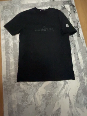 Svart Moncler t-shirt med logga - Svart t-shirt från Moncler med diskret logga på bröstet och Moncler-märke på ärmen. Klassisk passform och rund halsringning, perfekt för en stilren och enkel look. T-shirten är använd ett få tal gånger men är i perfekt skick😁