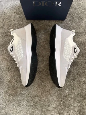 Vita sneakers från Dior med monogram - Snygga vita sneakers från Dior med gråa och vita detaljer samt tydligt Dior-monogram på sidorna. Skorna har svart sula med struktur, vita skosnören och meshpartier för extra andningsförmåga. Perfekta för dig som vill ha en exklusiv och trendig look.