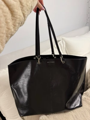 Svart shoppingväska från Ideal of Sweden - Stilren svart shoppingväska från Ideal of Sweden i glansigt konstläder. Väskan har dubbla axelband och metallfästen. Perfekt storlek för att få plats med allt du behöver. Enkel och klassisk design som passar till det mesta. Den är lite smutsig inne i väskan som man ser och axelbanden är sönder 