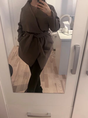 Mörkgrå omlottkappa med knytskärp - Snygg brun omlottkappa med knytskärp i midjan och avslappnad passform. Kappan har långa ärmar och en mjuk, slät yta som ger en stilren look. Perfekt för dig som gillar minimalistisk och trendig stil.
