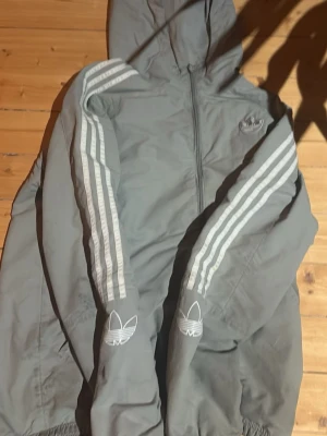 Grå Adidas vindjacka vintage  - Snygg grå vindjacka från Adidas med klassiska vita ränder längs ärmarna och huva. Jackan har dragkedja framtill, elastiska muddar och två sidofickor. Adidas-logga på bröstet och ärmarna ger en sportig vibe. Perfekt för dig som gillar streetwear och enkel stil.