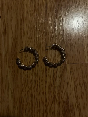 Guldiga tvinnade hoops örhängen - Säljer ett par chunky hoops i guldfärgad metall med tvinnad design. De är runda och har en cool, lite retro känsla som verkligen sticker ut. Perfekta för dig som gillar statementörhängen och vill addera lite extra bling till din look. De är ganska tunga. Kommer ej med sånna fästen 