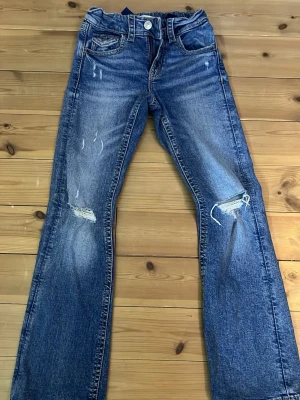Blå slitna bootcut jeans - Säljer ett par blå bootcut jeans med slitningar och hål på knäna. Jeansen har klassisk femficksdesign, knappgylf och ljusare tvättade partier för en cool vintage-look. Perfekta för dig som gillar en avslappnad och trendig stil.