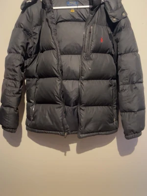 Svart dunjacka från Polo Ralph Lauren - Svart dunjacka från Polo Ralph Lauren med huva och dragkedja framtill. Jackan har en röd logga på bröstet och en bröstficka med dragkedja. Puffig modell som håller värmen under kalla dagar. Perfekt för vintern. Den har en liten skada på armen men det är inget som syns jätte mycket och det är enkelt att fixa.