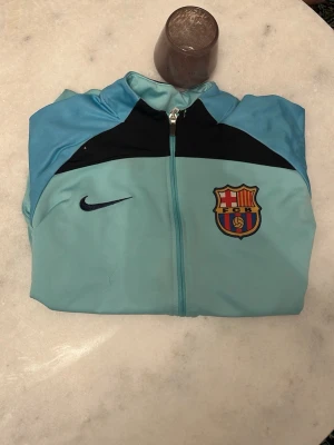 Nike FC Barcelona träningsjacka blå - Ljusblå träningsjacka från Nike med FC Barcelona klubbmärke på bröstet och svart Nike-logga. Jackan har hel dragkedja, ståkrage och svarta detaljer upptill samt grafiskt mönster på ärmarna. Perfekt för fotbollsträning eller som sportig vardagsstil. Har två fläckar man inte märker 