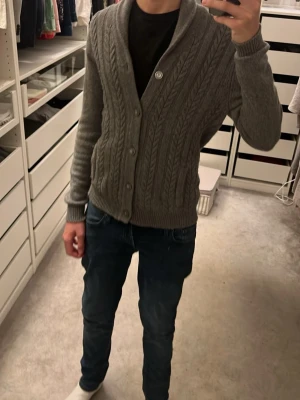 Grå stickad cardigan - Säljer en grå kabelstickad kofta med knappar framtill och bred krage. Koftan har ribbade muddar vid ärmslut och nederkant, och är perfekt för lager-på-lager. Passar dig som gillar klassisk och stilren design, hör av dej vid frågor o funderingar