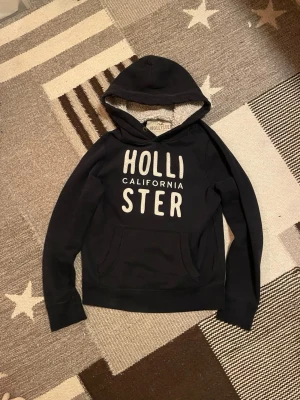 Hollister hoodie - Tja! Säljer denna riktigt goa hollister hoodien med vit ”Hollister California” logga på bröstet och med en riktigt snygg fodrad luva som tillför en cool karaktär till tröjan! Funkar lika bra under kalla vinterdagar som på sena  sommarkvällar och är superenkel att matcha!Tröjan är använd men har inga större defekter. Hör av dig vid minsta fundering! 😁🙌✅