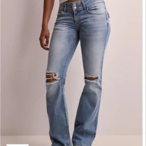 Bootcut ljusblå jeans med slitningar - Snygga ljusblå bootcut jeans med slitna hål över knäna och dubbla knappar i midjan. Jeansen har bakfickor med lock och knapp, samt klassisk femficksdesign. Perfekta för dig som gillar en avslappnad och trendig look.