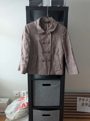 Beige kavajjacka från Caroll - Snygg beige kavajjacka från Caroll med dubbelknäppning och stora knappar framtill. Jackan har en mjuk krage, raka fickor och är tillverkad i ett linneliknande material. Perfekt för dig som gillar klassisk stil med en twist.