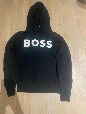 Boss hoodie - Snygg svart hoodie från BOSS med stor vit logga på bröstet. Klassisk huva med dragsko och magficka framtill. Tillverkad i mjukt material som känns skönt mot huden. Pefekt för en snygg outfit till vardagen eller om du vill lyxa till det på helgen. Passar dig mellan 170-180 cm.