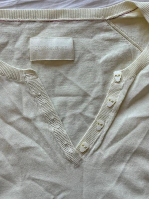 Ljusgul topp med dödskalleknappar - Ljusgul topp från Zadig & Voltaire med korta ärmar och ribbade kanter. V-ringning med unika  dödskalleknappar längs öppningen framtill. 