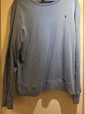 Ljusblå sweatshirt från Polo Ralph Lauren - Ljusblå sweatshirt från Polo Ralph Lauren med klassisk rund halsringning och diskret broderad logga på bröstet. Tröjan har långa ärmar och ribbade muddar vid ärmslut och nederkant. Perfekt för en avslappnad och stilren look.